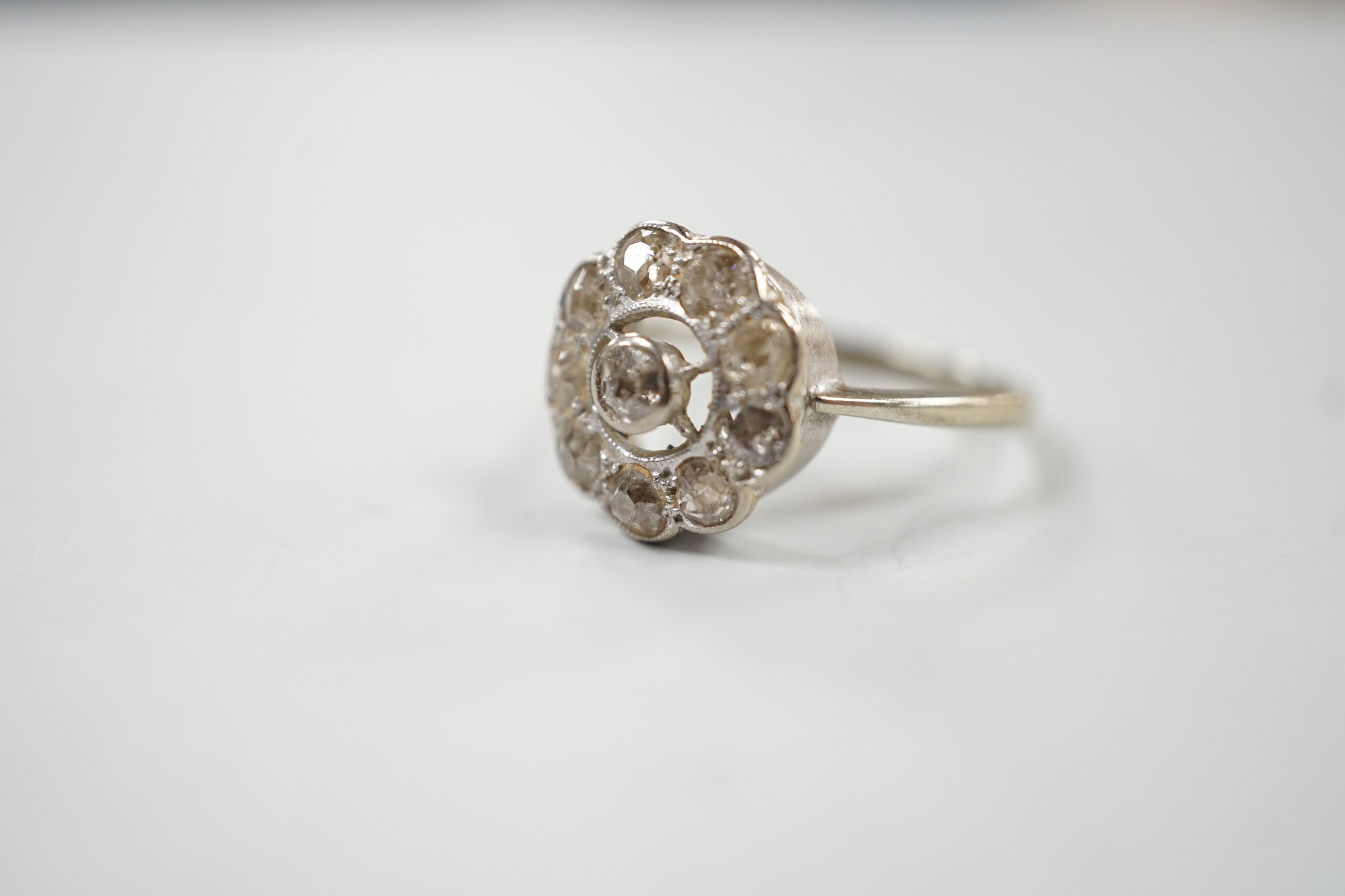 A 1920's white metal and ten stone millegrain set diamond flower head cluster ring, size P/Q, gross weight 3 grams.
